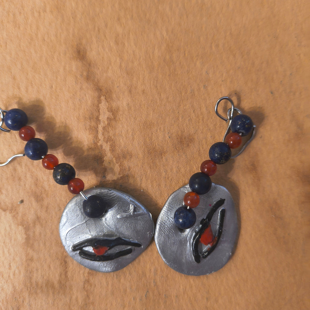 Pair of silver earrings with black eye hieroglyphics. Each earring has three purple beads and three red beads. Sitting on a brown surface. 