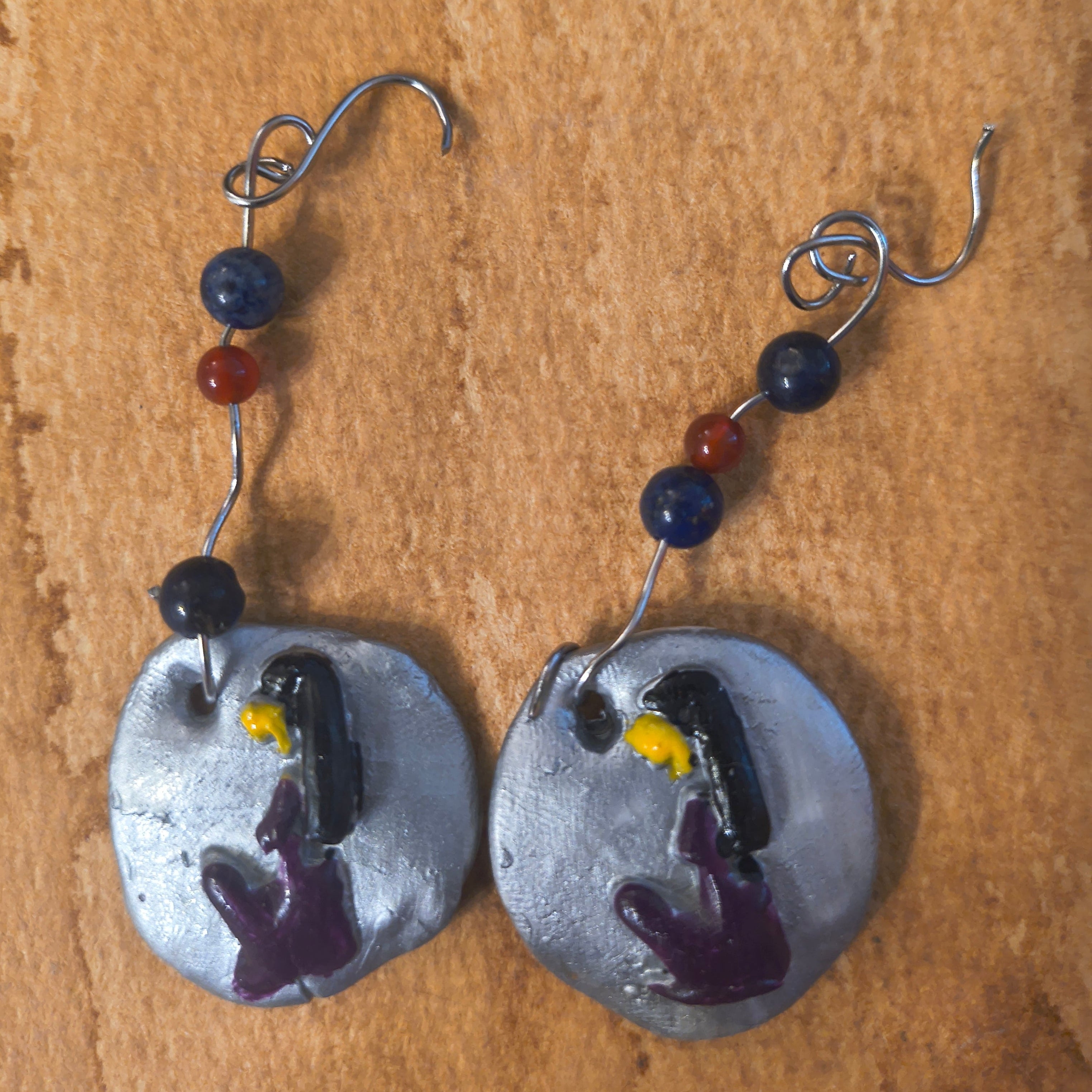 Pair of silver earrings with purple hieroglyphics of a woman. Earrings have two purple beads and one red bead. Sitting on a brown surface.
