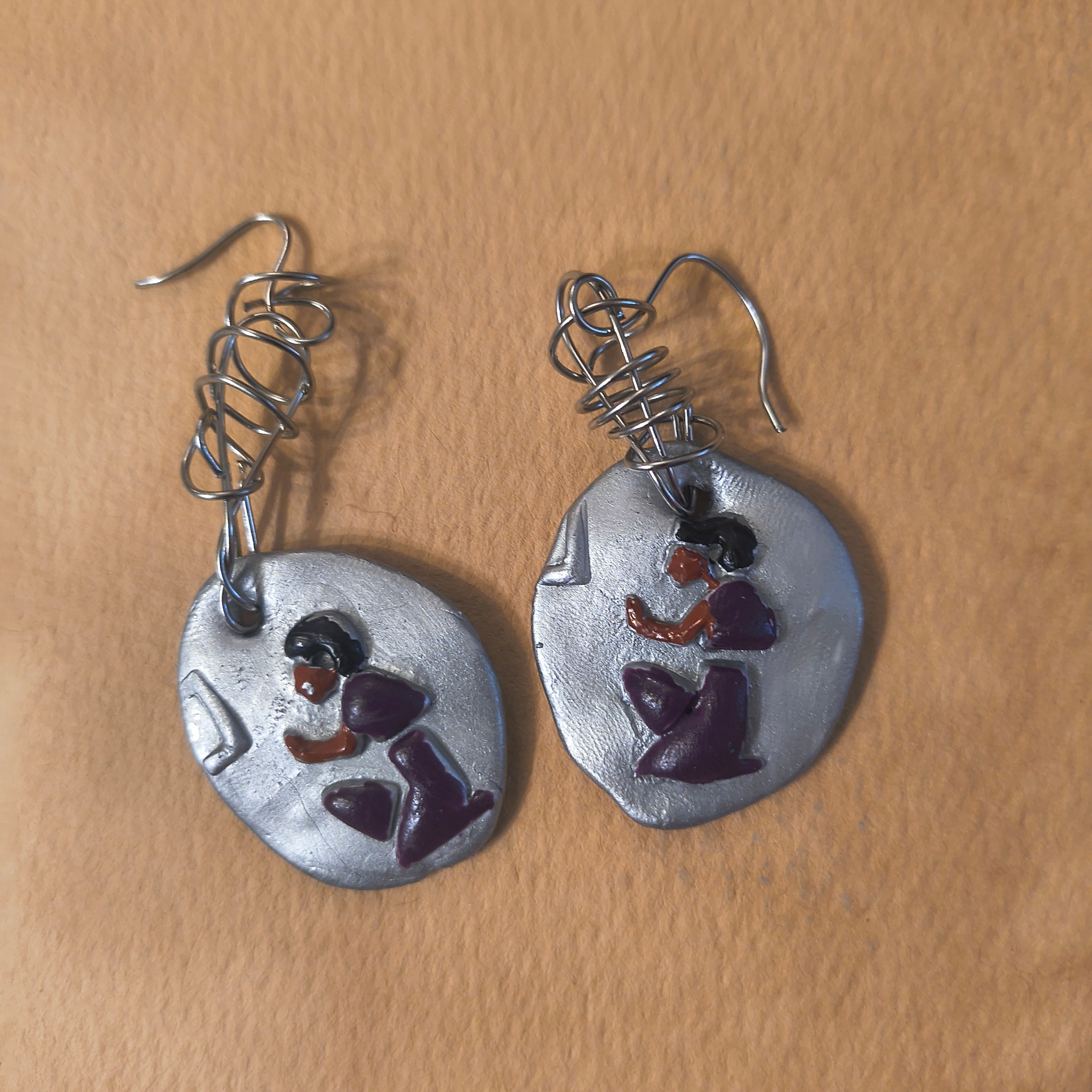 Pair of silver earrings with purple hieroglyphics of a man. Sitting on a brown background. 